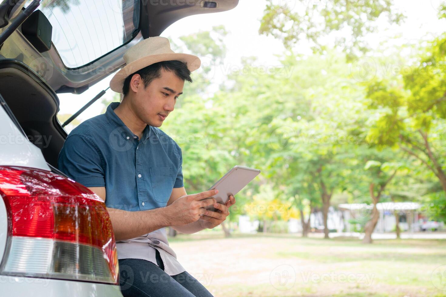 Asian man sitting back of car and holding tables check location and find place to travel. Happy road trip and vacation in summer in the countryside. Concept of journey on leisure, road trip vacation photo