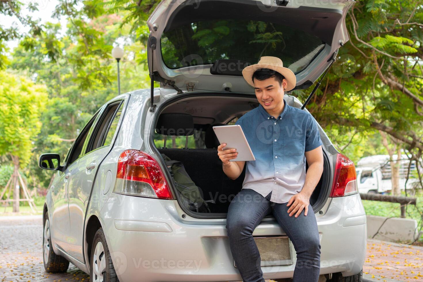 Asian man sitting back of car and holding tables check location and find place to travel. Happy road trip and vacation in summer in the countryside. Concept of journey on leisure, road trip vacation photo