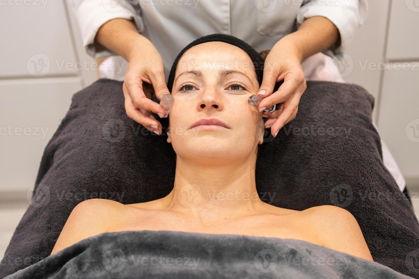 Crop beautician massaging cheeks of client with stones photo