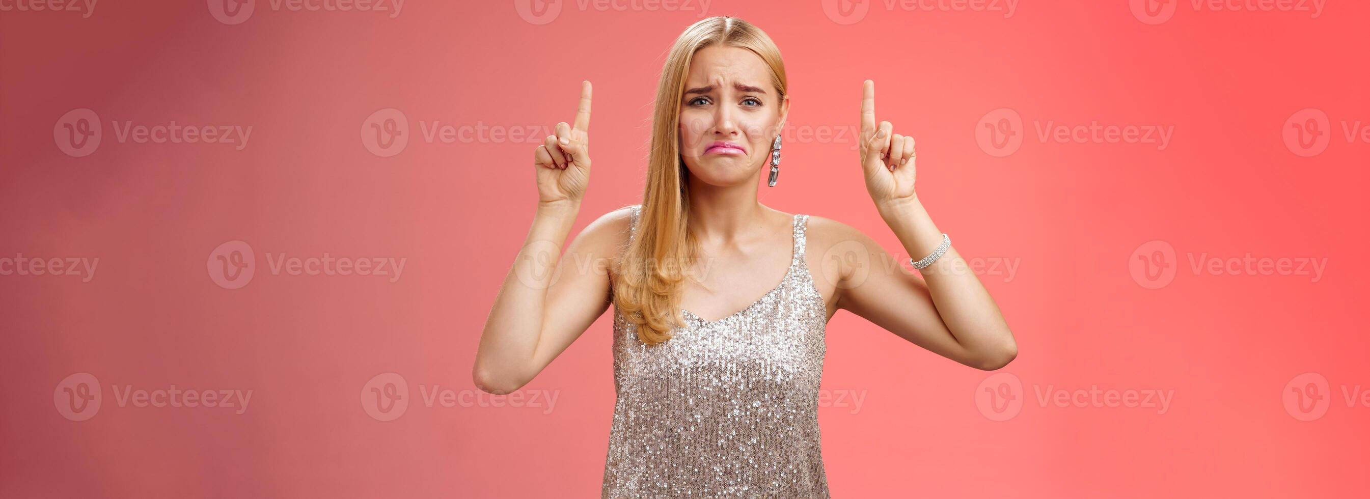 Upset whining immature spoiled blond rich girl in silver glittering dress pouting frowning gonna cry pointing up regret jealousy begging buy expensive shoes, standing red background displeased photo