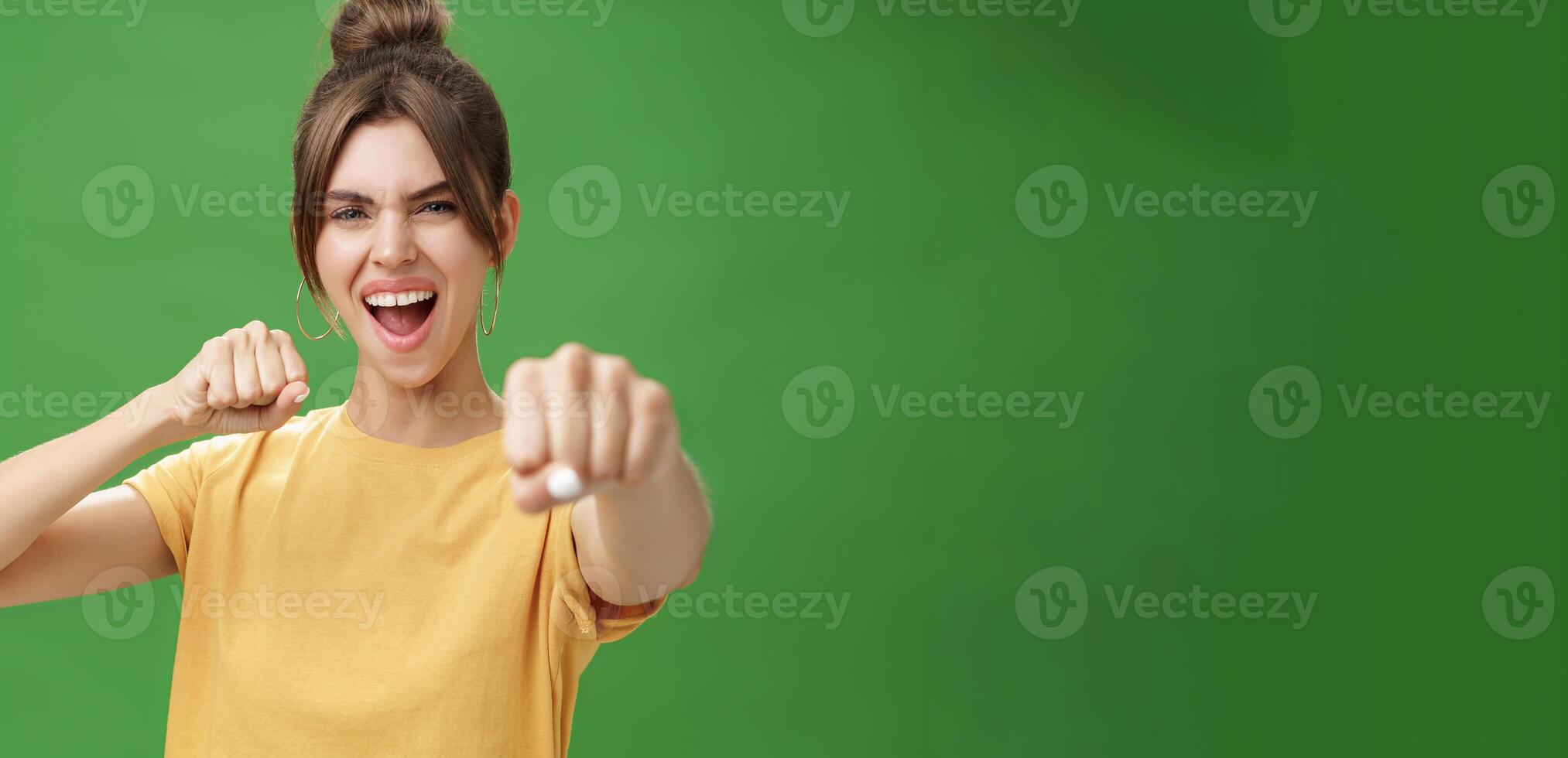 Cute female rebel in yellow t-shirt with gap teeth pulling fist towards camera as if showing fighting skills yelling daring and excited standing over green background smiling acting like boxer photo