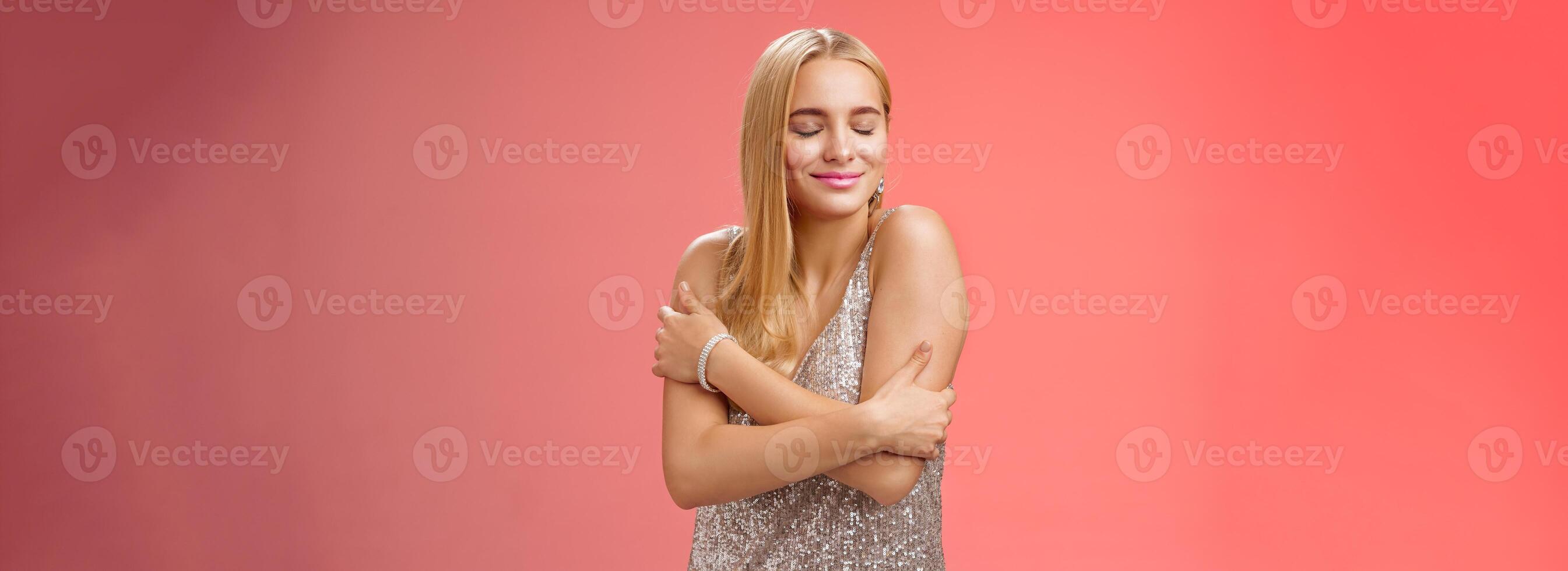 Tender feminine elegant blond young woman in stylish silver glittering dress close eyes dreamy smiling touching herself cuddle embrace hands crossed body remember heartwarming passionate feelings photo