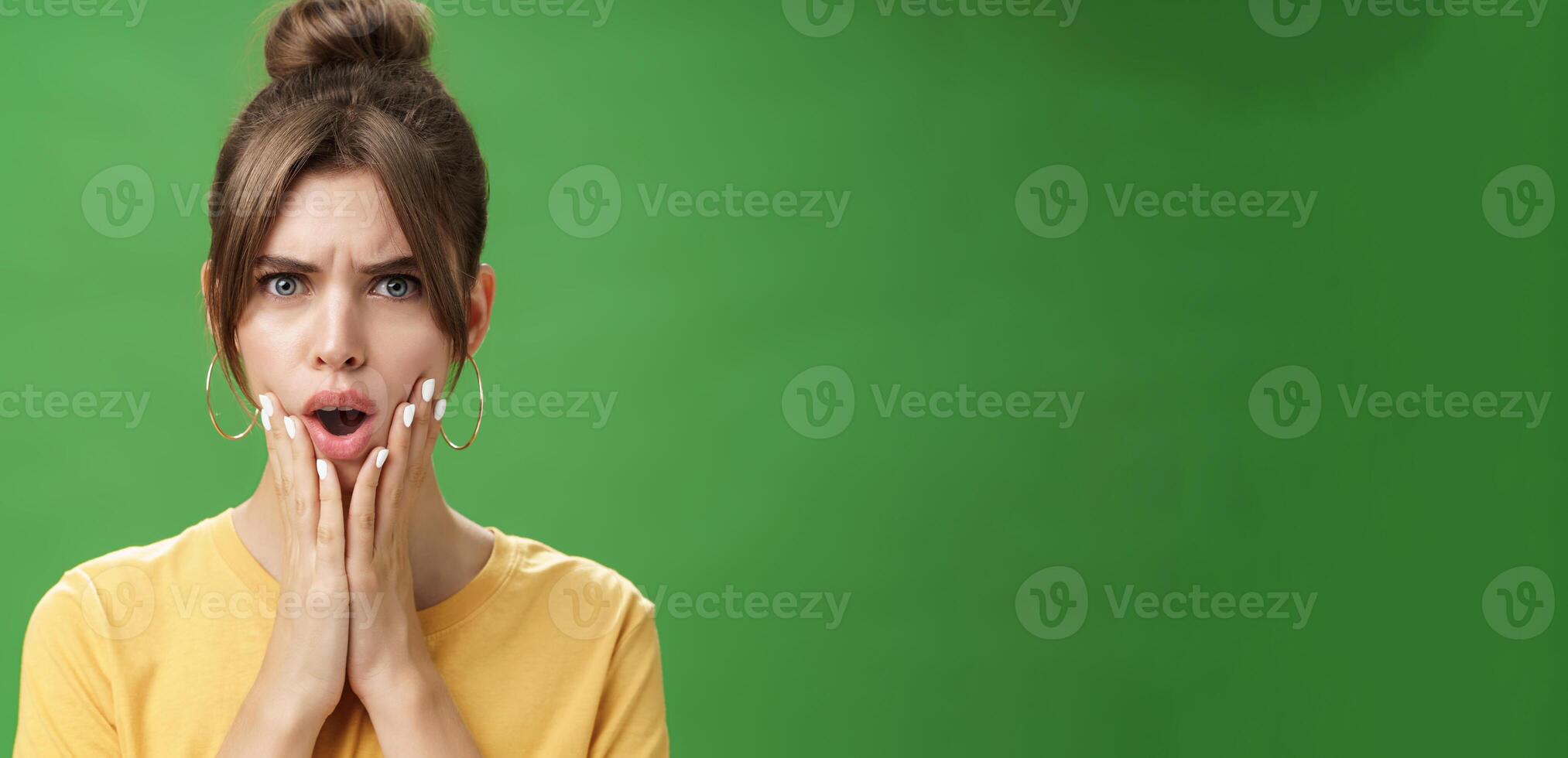 Shocked woman reacting to pimple looking disturbed and displeased in mirror holding hands pressed to cheeks open mouth and frowning being disappointed and upset over green background photo