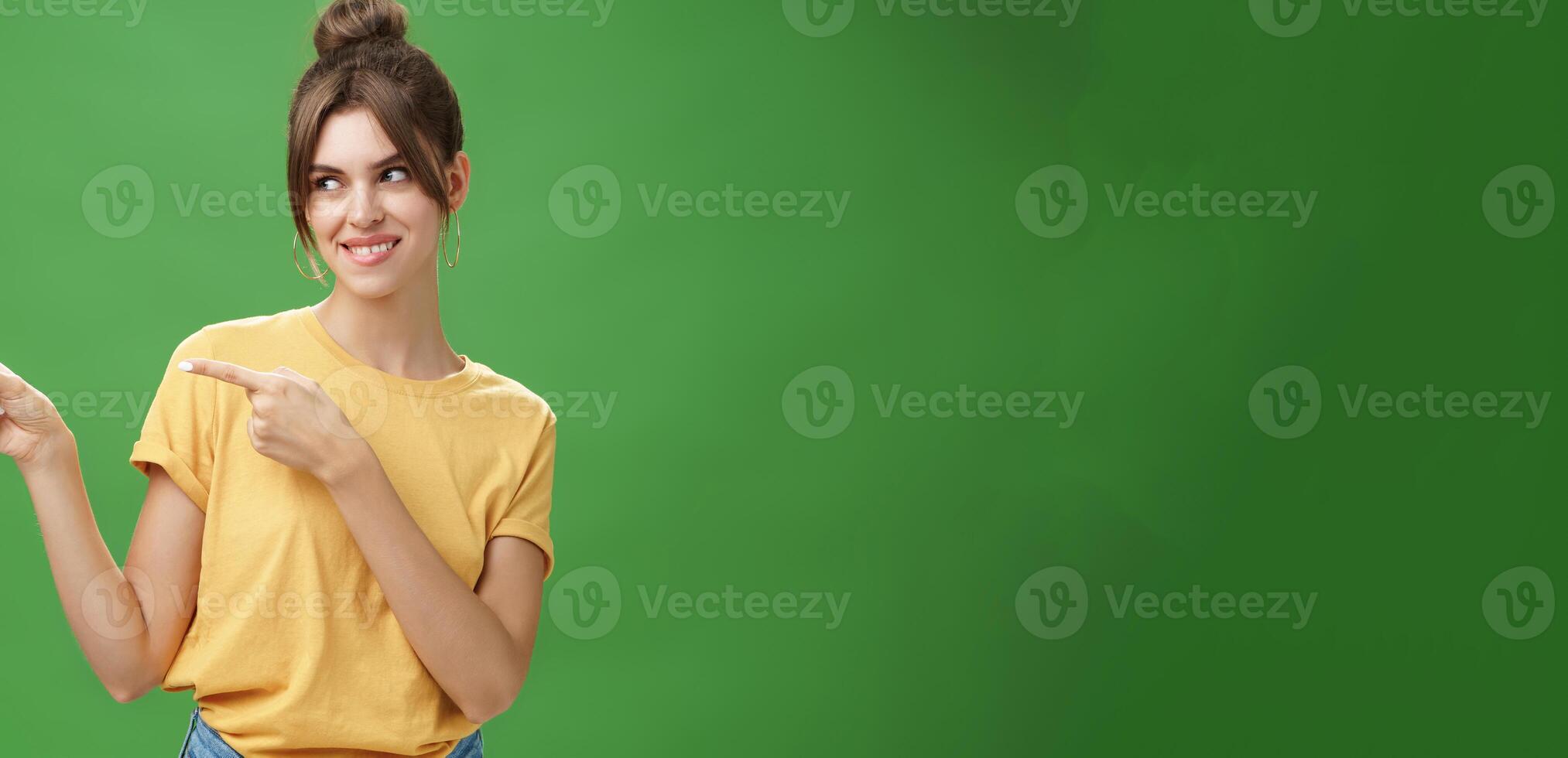 Waist-up shot of charismatic happy and carefree charming woman in yellow t-shirt pointing and looking left enthusiastic and pleased smiling cheerfully posing against green background delighted photo