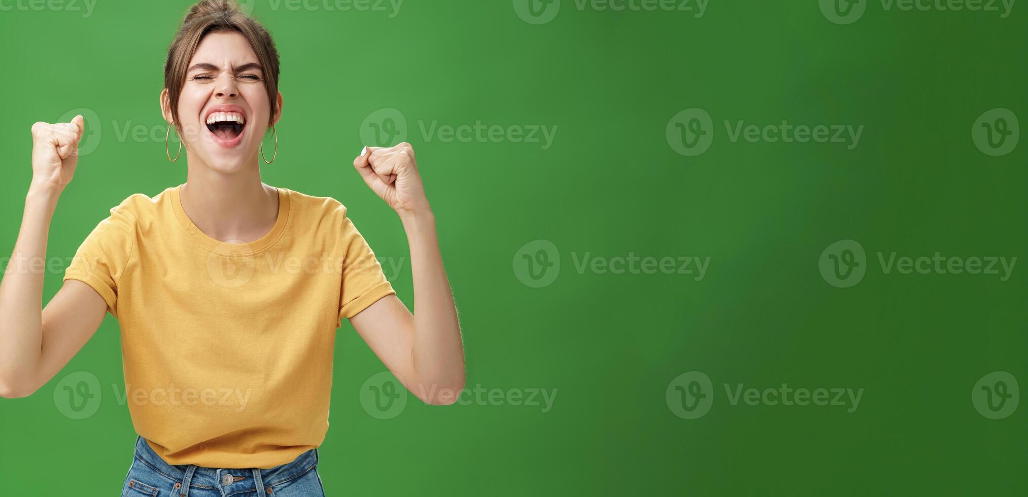 Waist-up shot of charismatic energized and excited female in yellow t-shirt closing eyes yelling from joy and happiness raising hands in cheer celebrating successful news over green background photo