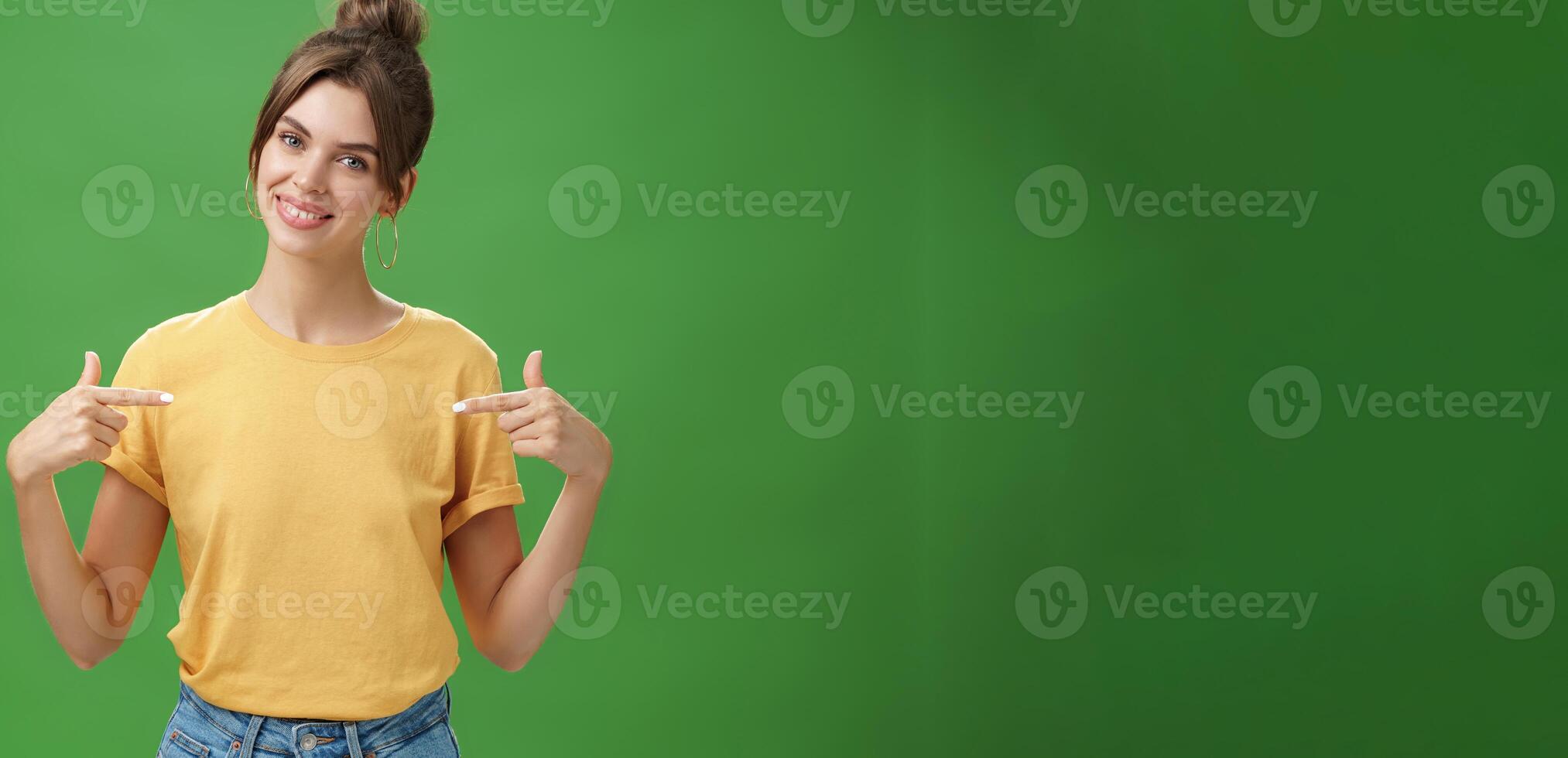 Calm and polite friendly-looking skillful young female coworker in yellow t-shirt pointing at herself self-assured tilting head and smiling at camera wanting participate in event over green background photo