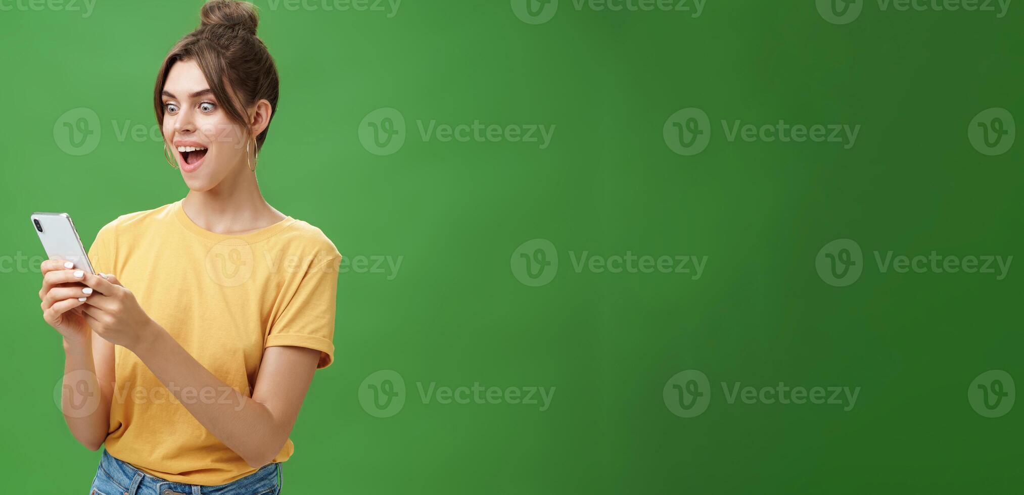 Woman reading surprising satisfying message in smartphone opening mouth from excitement, smiling amazed looking astonished at cellphone screen posing against green background in casual yellow t-shirt photo