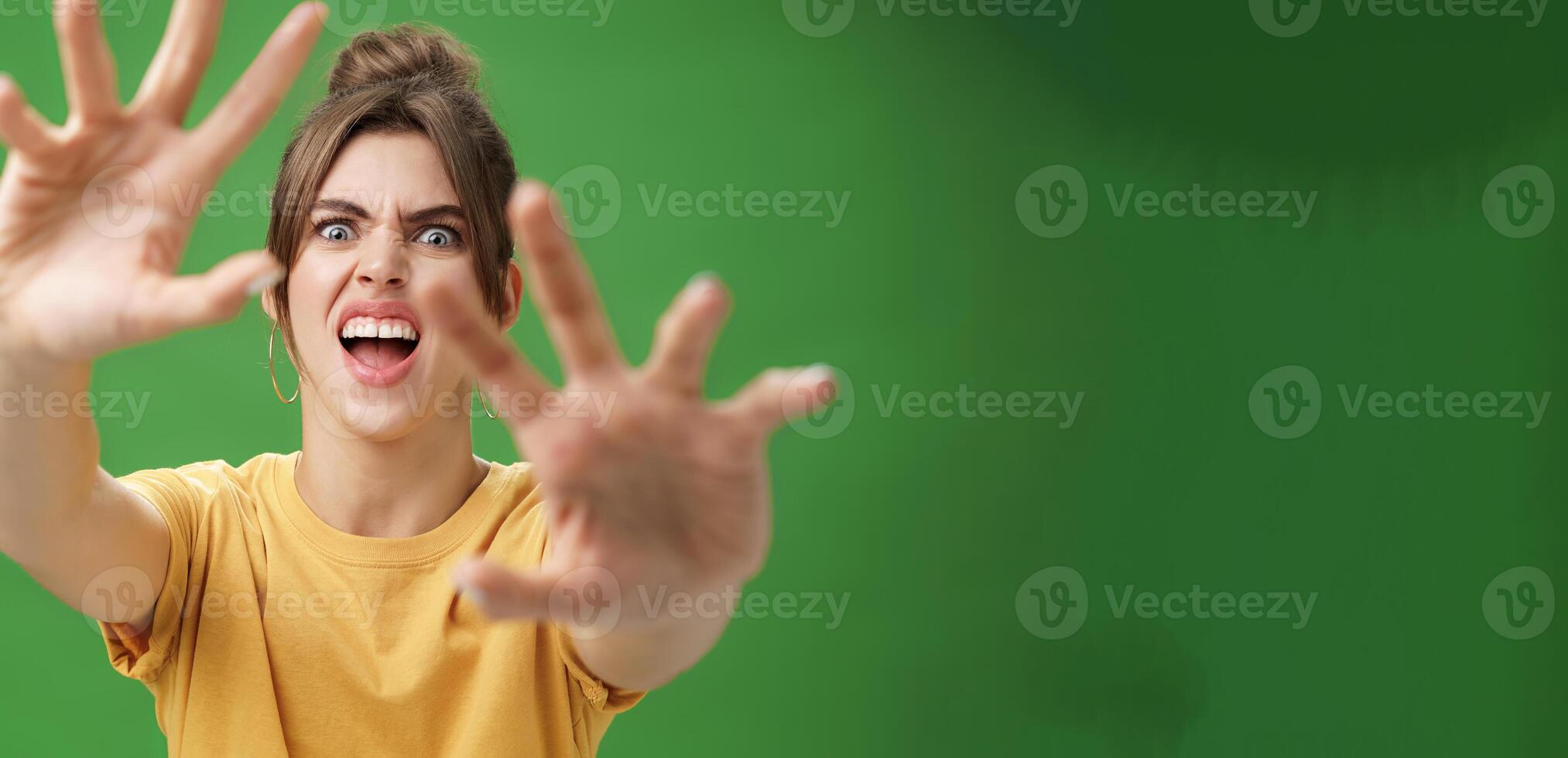 Funny emotive woman making fun face pulling hands forward to attack of grab something frowning opening mouth wanting cover face from camera, grimacing over green background photo