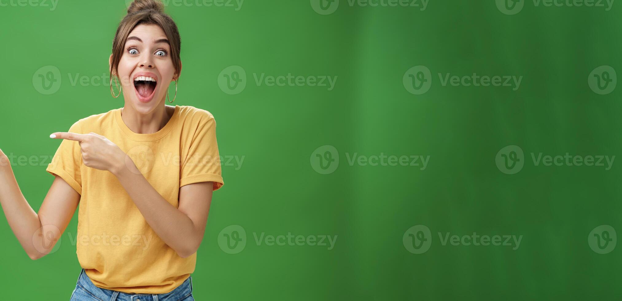 Waist-up shot of enthusiastic and charismatic impressed happy girl laughing and smiling broadly pointing left at funny hilarious thing reacting surprised and entertained, posing over green background photo