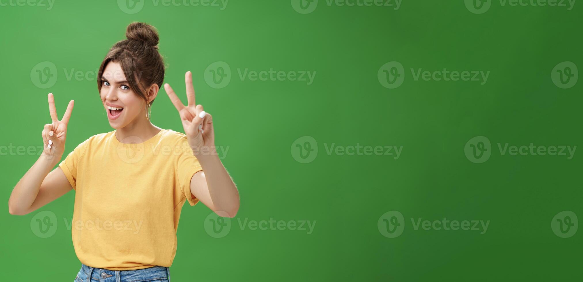 Indoor shot of enthusiastic excited and happy daring girl with combed hair tattoo and cute diasdema showing peace signs bending backwards standing in cool energized pose over green background photo