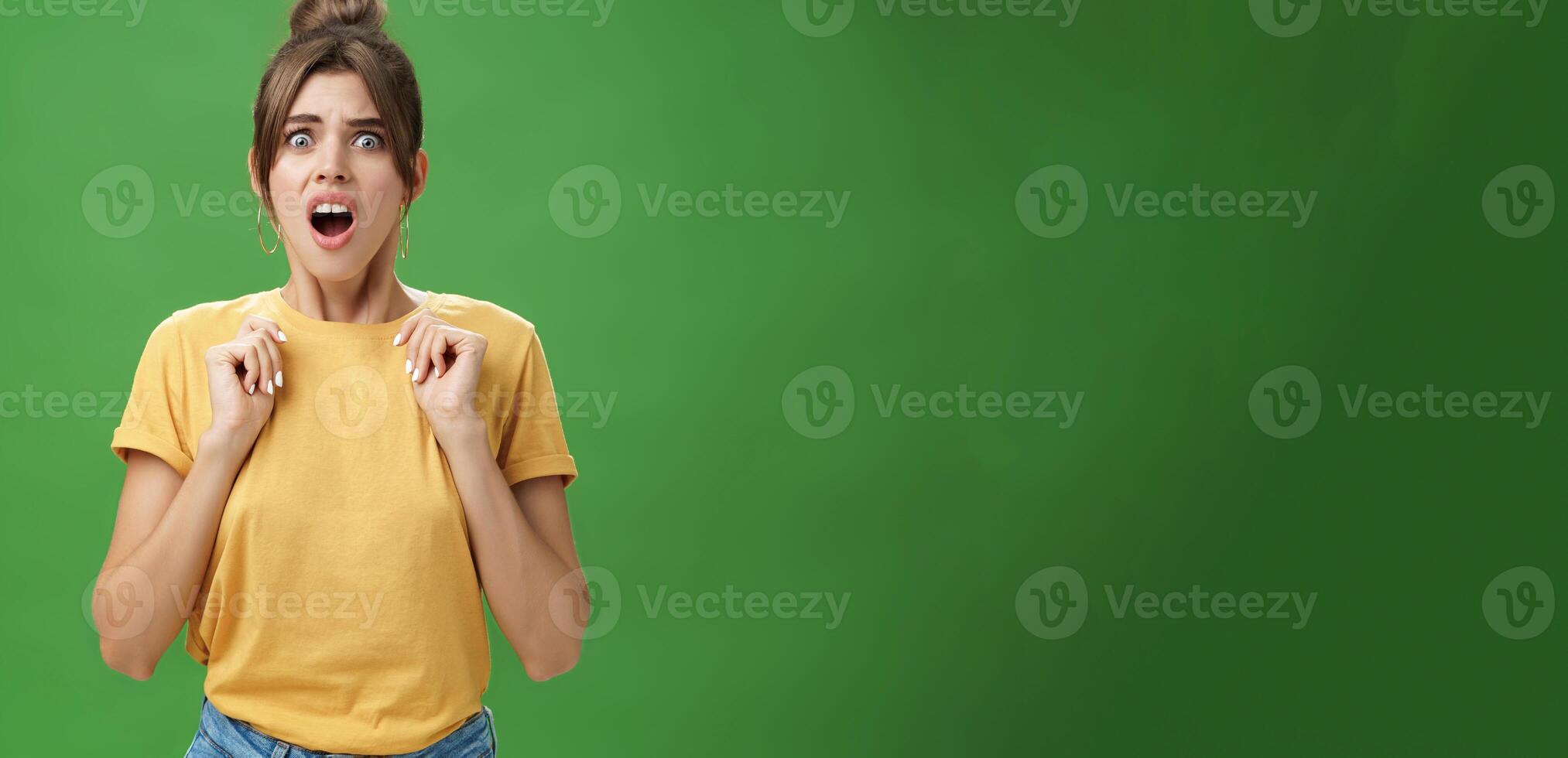 Shocked feminine glamour woman with combed hair in yellow t-shirt frowning gasping with opened mouth holding palms pressed to chest from aversion and disgust looking at creepy thing over green wall photo