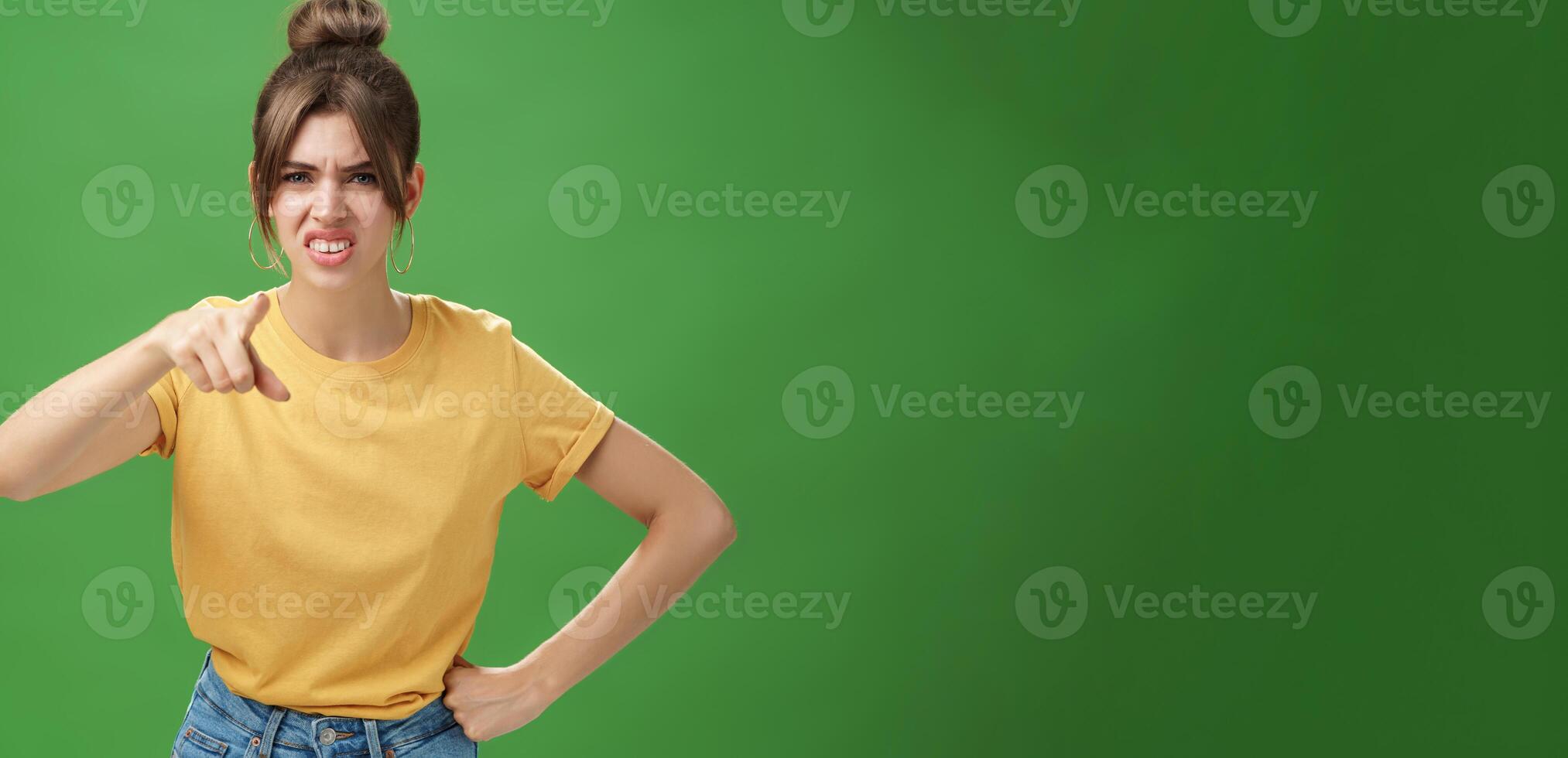 Portrait of arrogant impolite young woman in yellow t-shirt pointing at camera squinting and grimacing from disdain and scorn standing unimpressed judging against green background photo