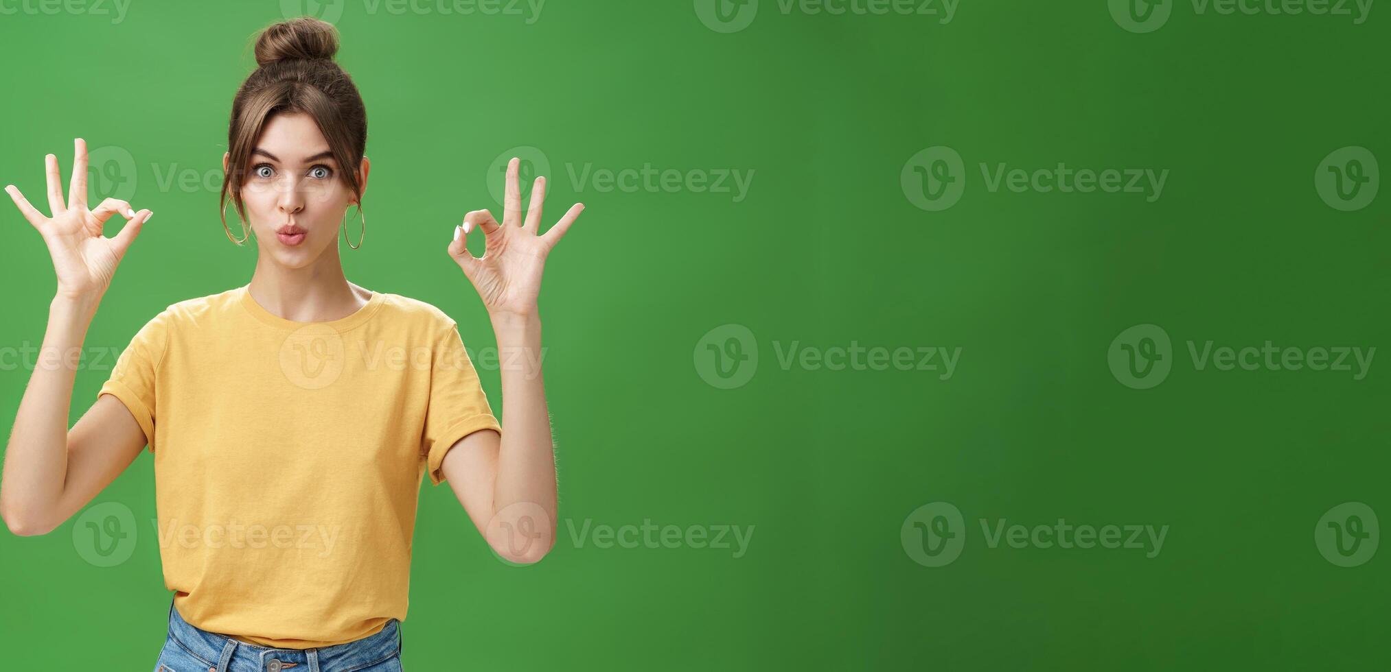 Portrait of silly cute and beautiful caucasian woman with combed hair in round earrings and yellow t-shirt showing okay, no problem gesture folding lips, reaffirming boss job goes well over green wall photo