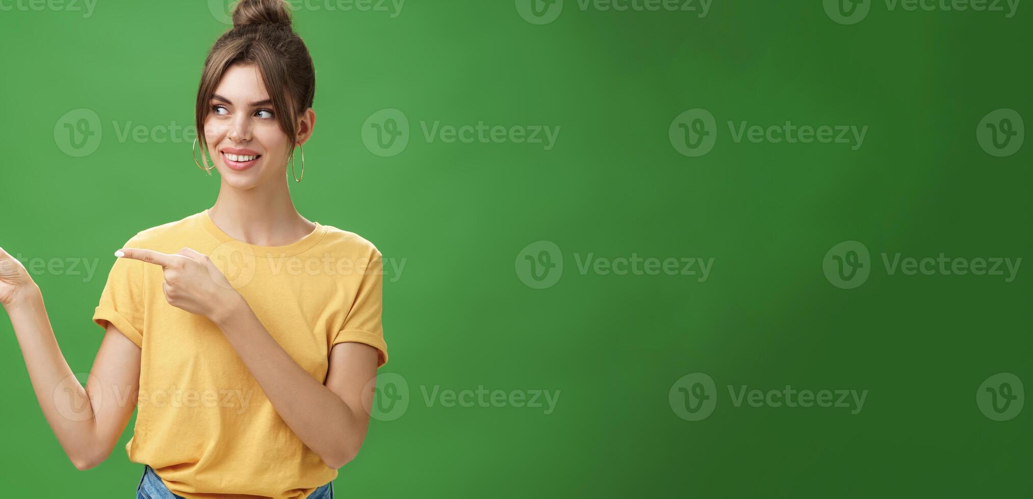 Waist-up shot of good-looking friendly and feminine female coworker in yellow t-shirt showing newbie office pointing and looking left with cute happy smile posing against green background photo