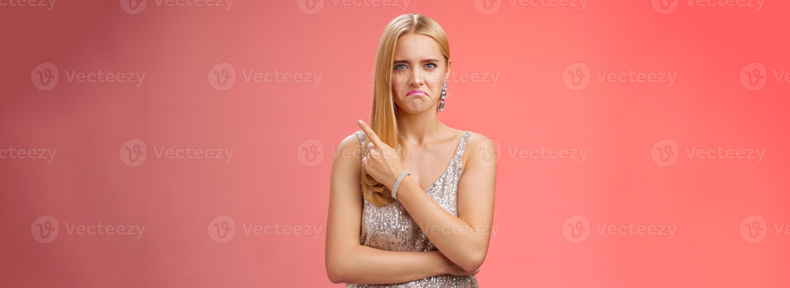 Jealous displeased angry young revengeful blond ex-girlfriend in luxurious silver shiny dress frowning pouting pointing upper right corner displeased pissed standing fed up upset red background photo