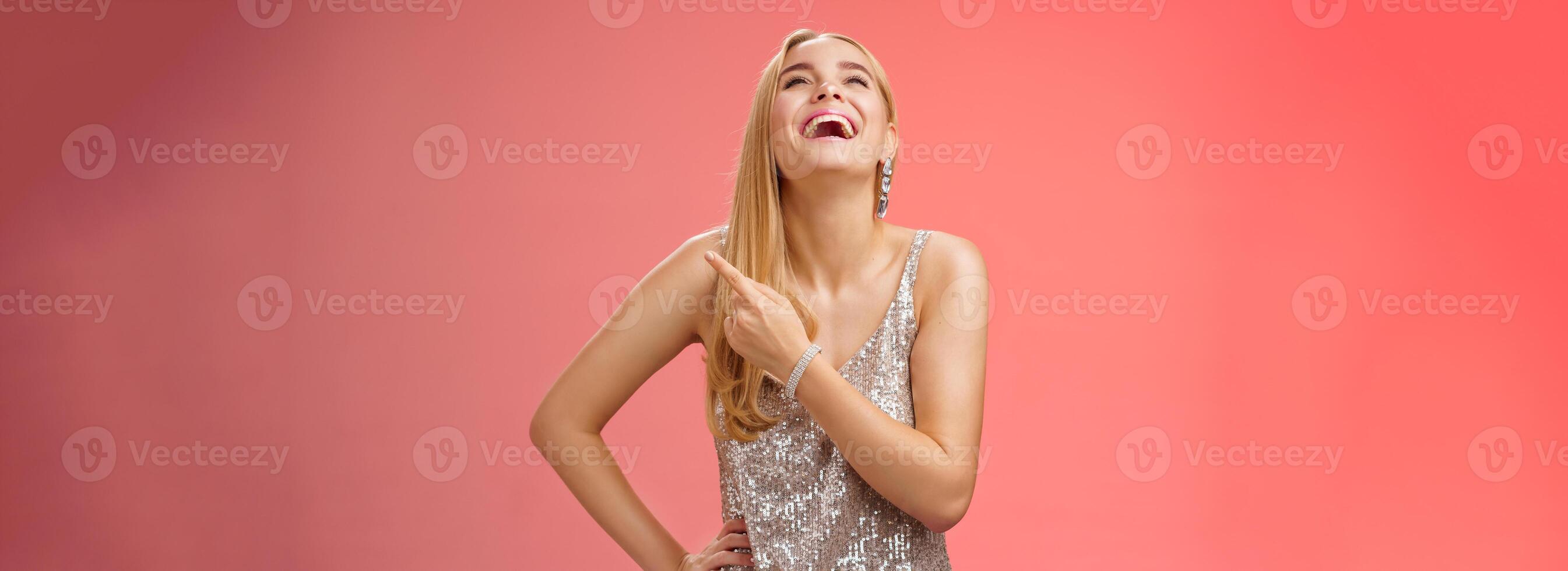 Fabulous carefree attractive blond woman in silver evening party dress laughing out loud have fun raise hand joyfully up pointing right index finger enjoying awesome sense humour, red background photo