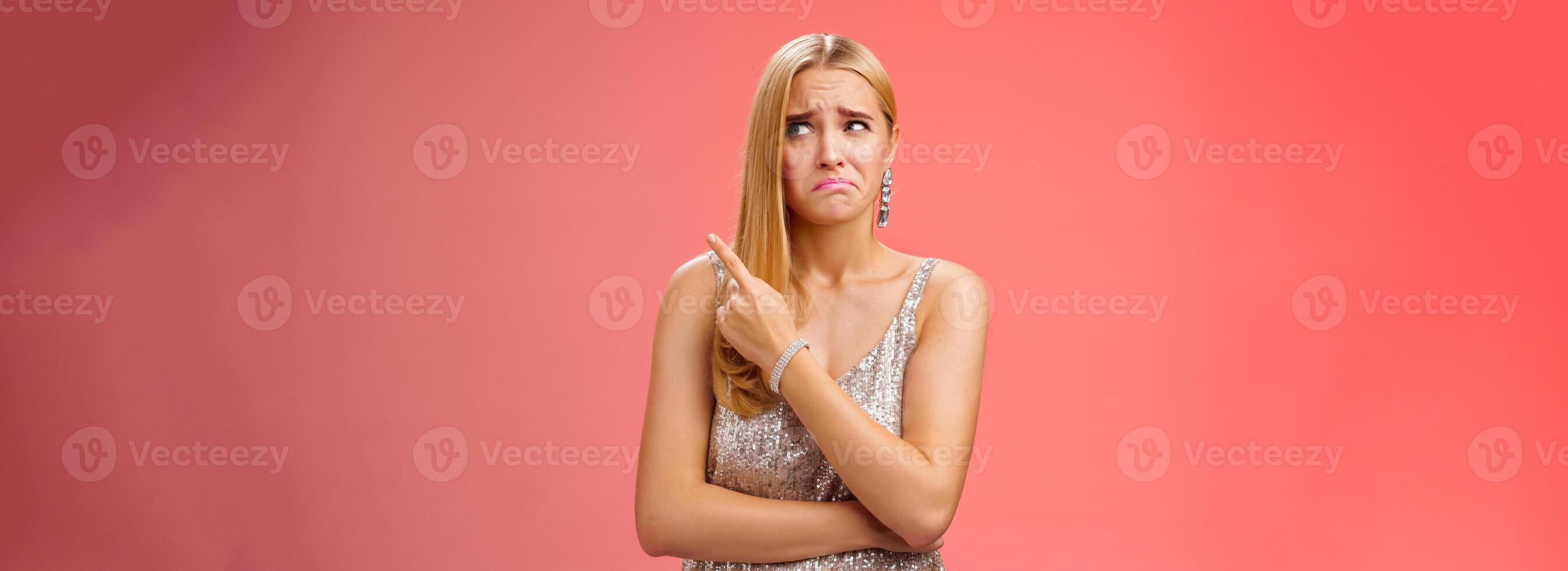 Upset doubtful unsure cute blond funny glamour girl in silver evening dress cringing grimacing hesitant pointing looking upper right corner suspicious, uncertain standing red background photo