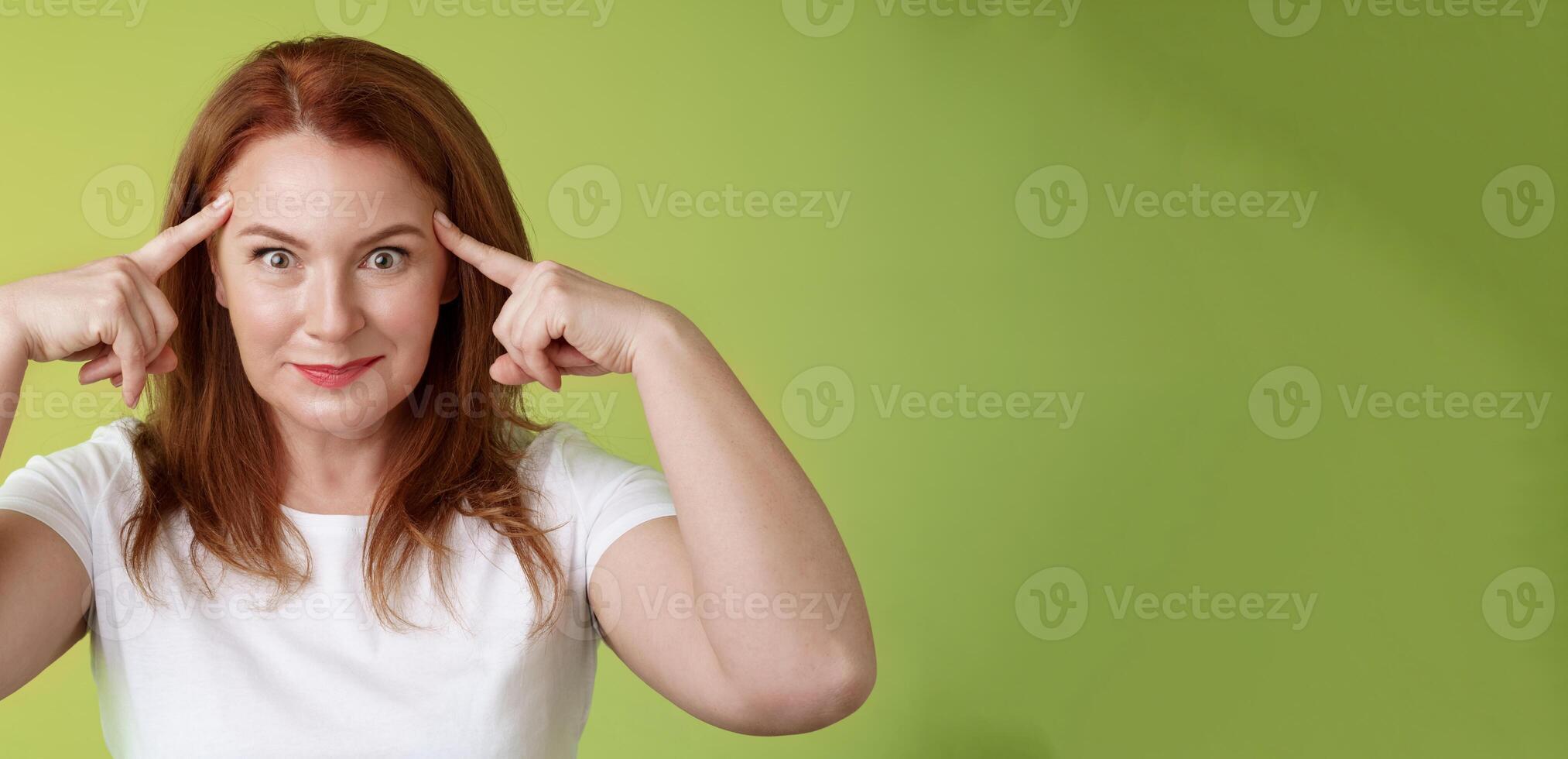 Woman playfully staring funny control your mind. Silly redhead middle-aged female touch temples popping eyes smiling delighted read thoughts trying guess intrigued what thinking green background photo