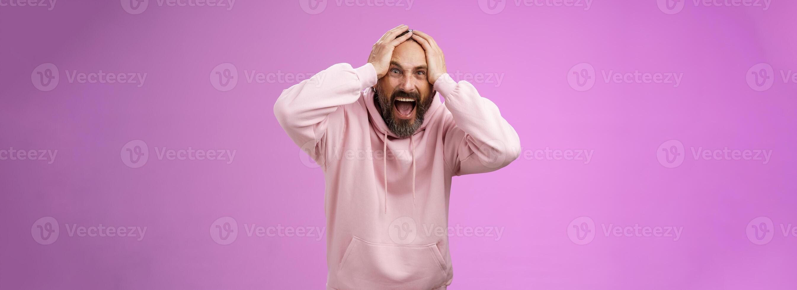 Outraged furious mad adult man bearded yelling shocked despair hatefully looking camera insane holding hands head frowning shouting losing bet disappointed very upset standing purple background photo