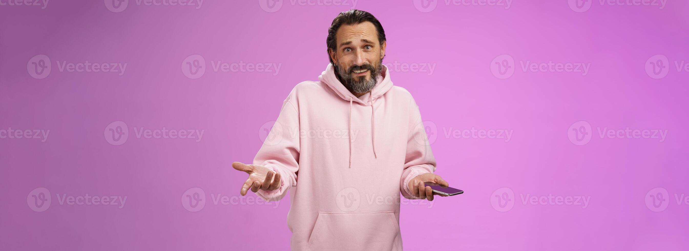 So what. Confused questioned adult bearded stylish guy in pink hoodie shrugging hands spread sideways dismay holding smartphone look clueless unaware camera not know how deal annoying phone calls photo