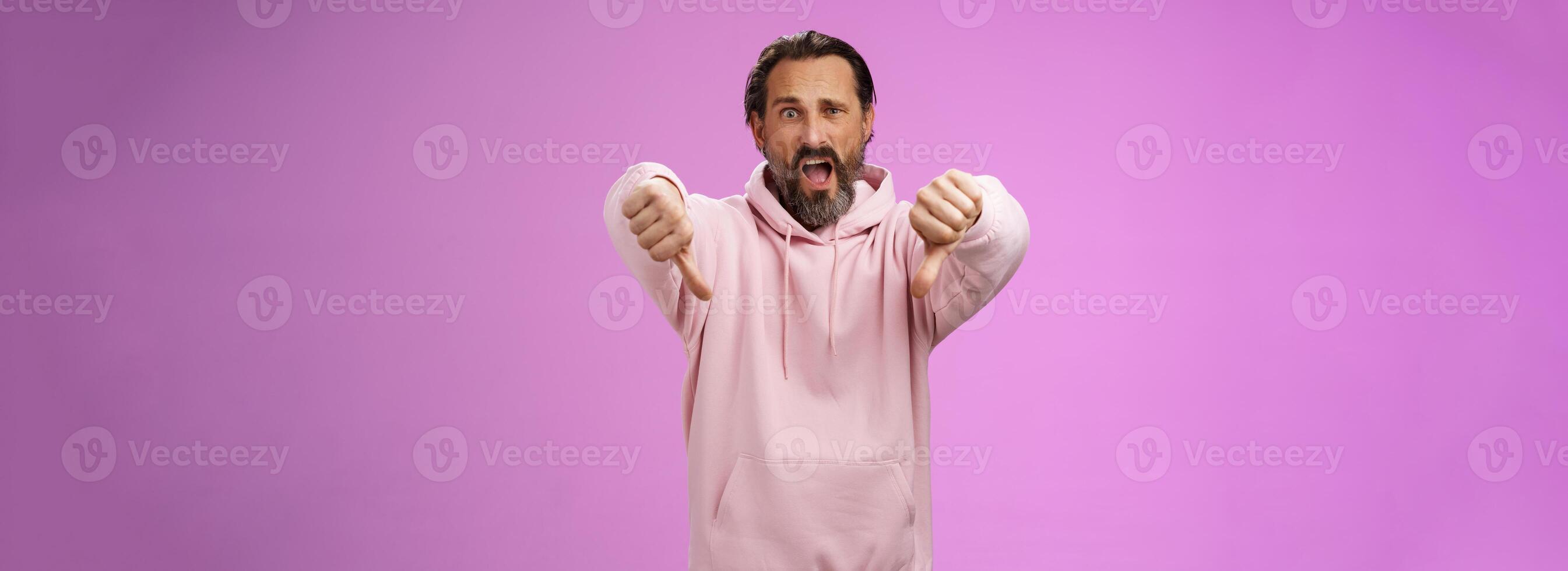 Extremely disappointed furious male customer swearing show thumbs down impulsive gesture yelling displeased pissed express dislike disgust look judgemental bothered, standing purple background photo