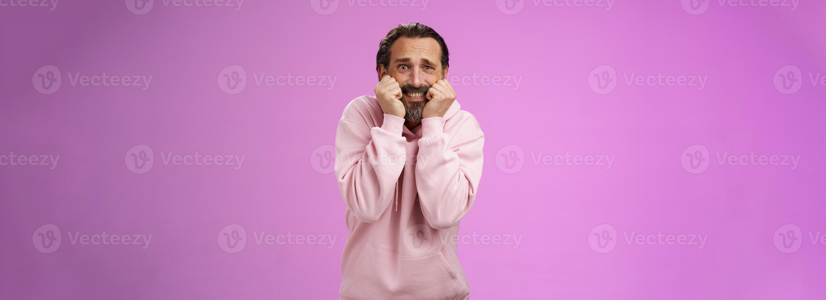 Scared insecure silly adult bearded man grey hair in pink hoodie press palms mouth bite fingers clench teeth shocked frightened widen eyes terrified standing stupor horrified, purple background photo