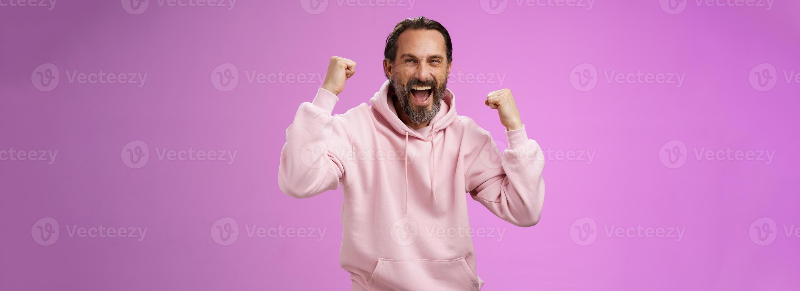 Cheerful supportive manly mature adult bearded guy fan yelling raising clenched fists triumphing team scored goal celebrating standing pleased shouting achieving success, posing purple background photo