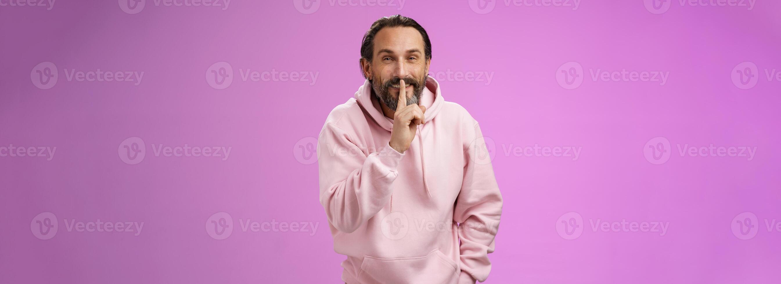 Lips sealed secret. Portrait alluring cheeky stylish adult bearded guy in pink hoodie asking keep quiet show shush shh gesture index finger pressed mouth smiling devious smug face, have idea photo