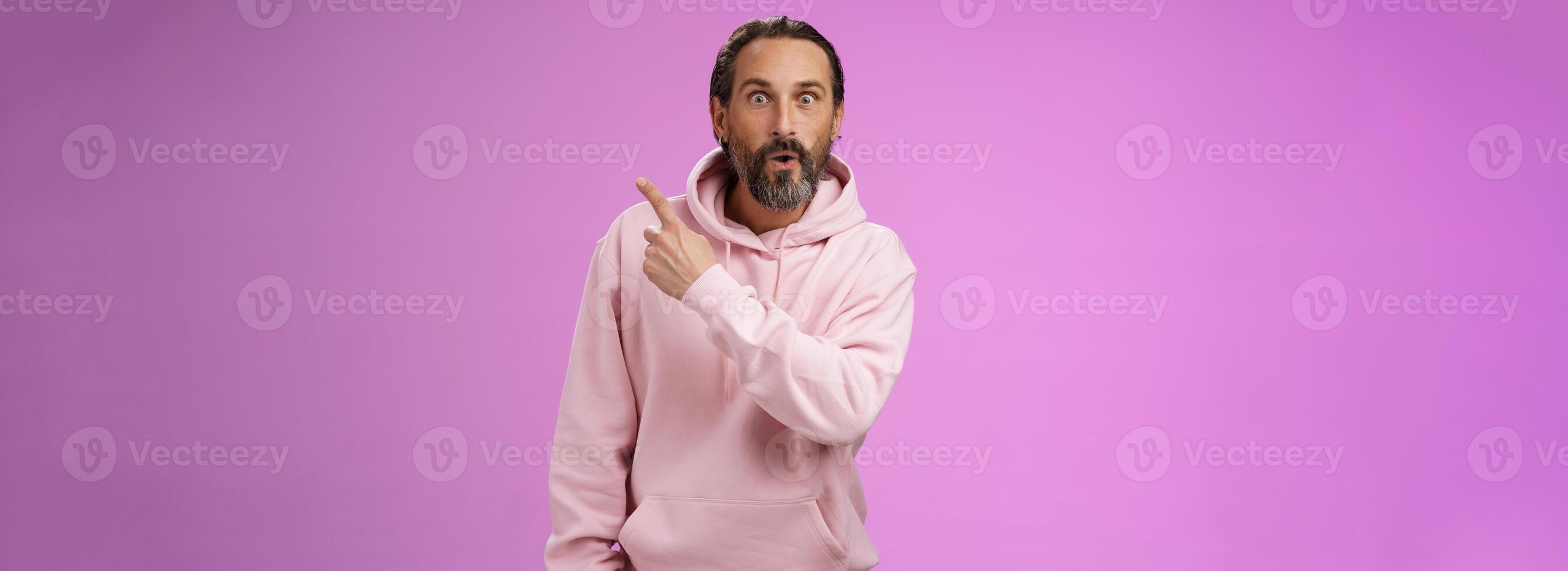 Amused handsome hipster mature 40s bearded caucasian man grey hair wearing trendy pink hoodie fold lips amazed wow pointing upper right corner widen eyes surprised check out awesome product photo