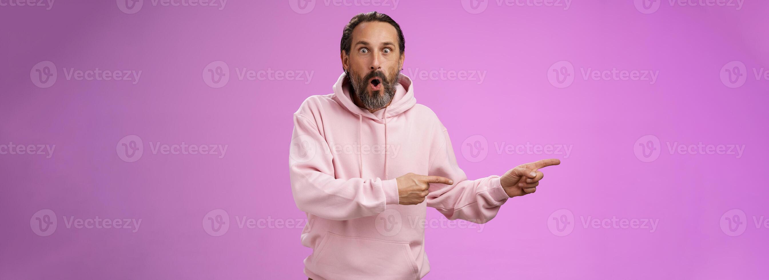 Astonished fascinated curious mature bearded man with grey hair in pink hoodie folding lips amazed pointing left index fingers widen eyes camera impressed thrilled wanna go check out cool promo photo