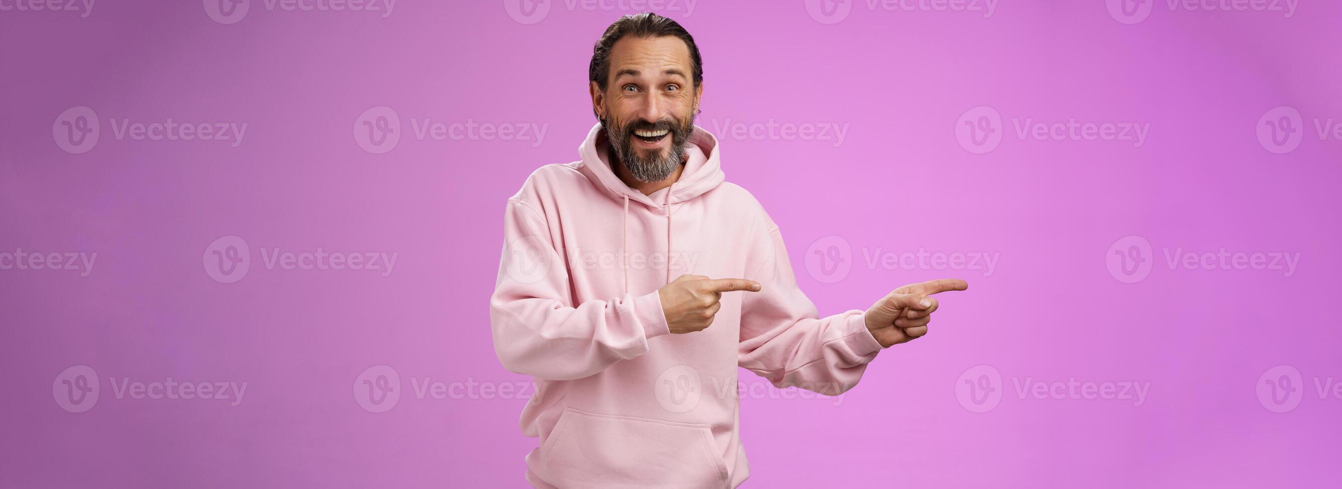 Charismatic happy good-looking mature 50s man wearing stylish hipster hoodie smiling excited pointing left index fingers impressed awesome interesting proposal standing pleased purple background photo