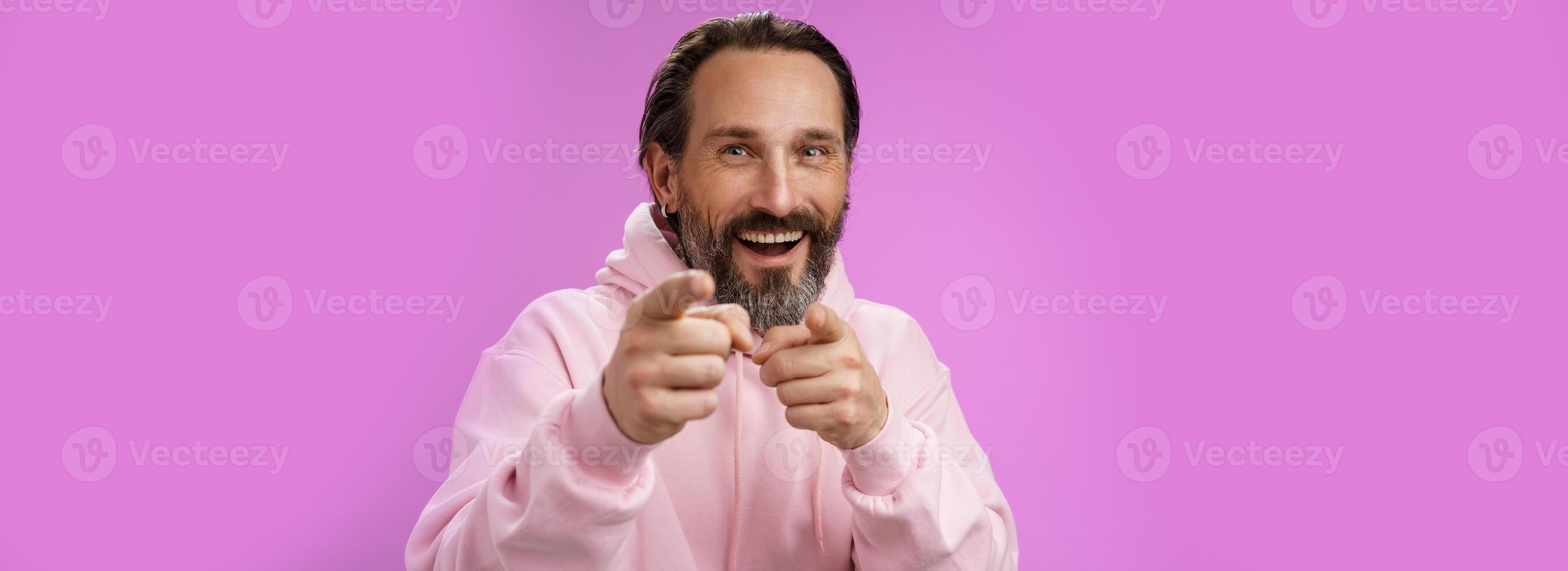 Funny amused carefree happy adult handsome man pranking friend fool around pointing camera index fingers greeting choosing you laughing joyfully having fu, standing purple background rejoicing photo