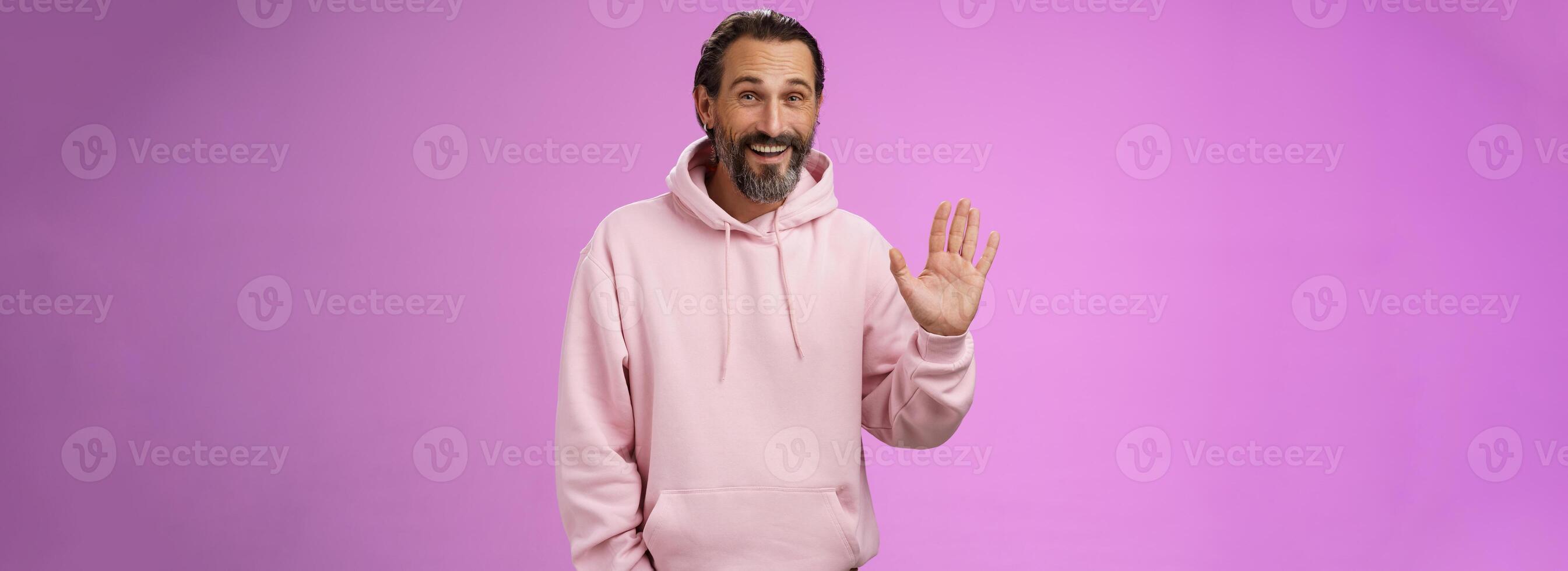 Cheeky charismatic funny happy smiling mature man bearded grey hair in pink hoodie waving palm hello nice meet you greetng gesture welcoming guests say hi standing purple background friendly relaxed photo