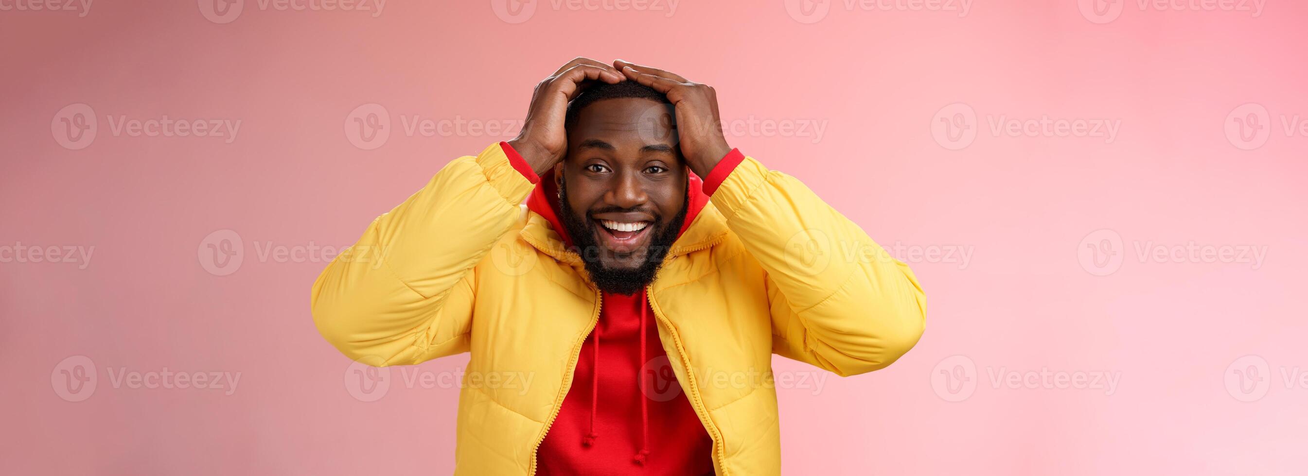 Waist-up surprised happy lucky young african-american bearded male in yellow jacket holding hands head smiling impressed joyfully receive awesome proposal dream come true, pink background photo