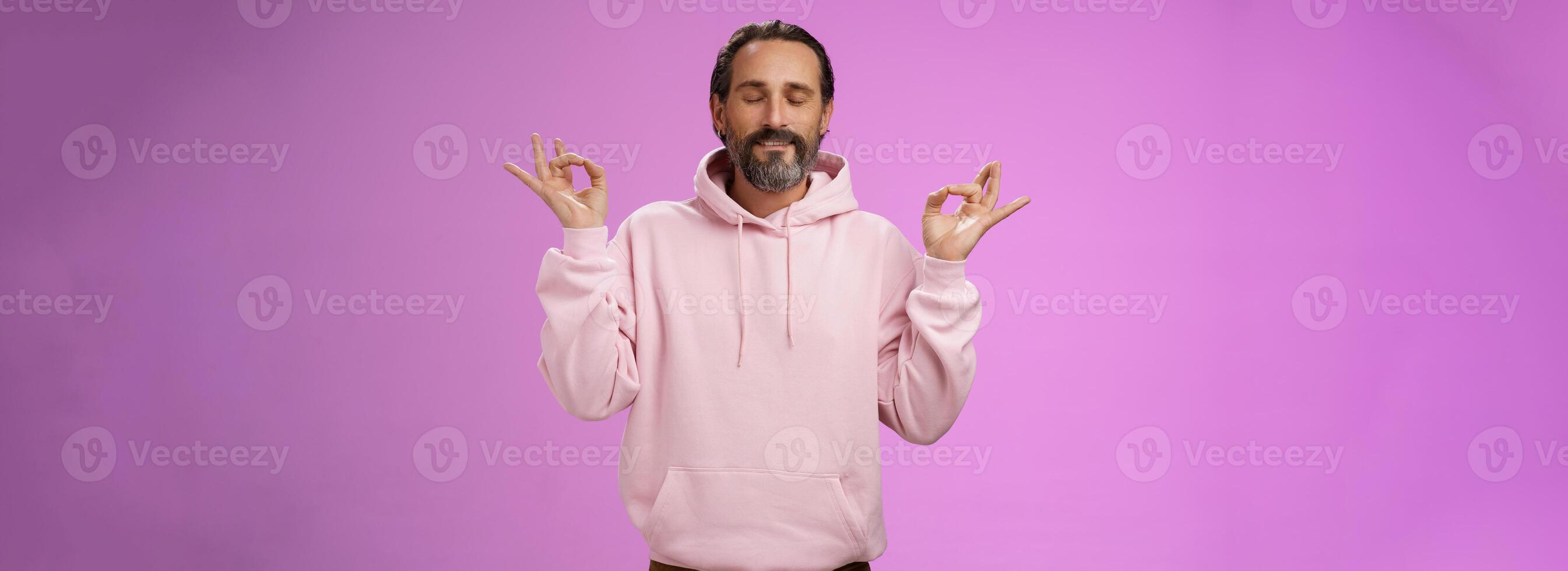 Peaceful charming hipster old man wearing cool pink hoodie close eyes breathing practice release stress meditating standing relaxed happy yoga meditation pose nirvana gesture, purple background photo