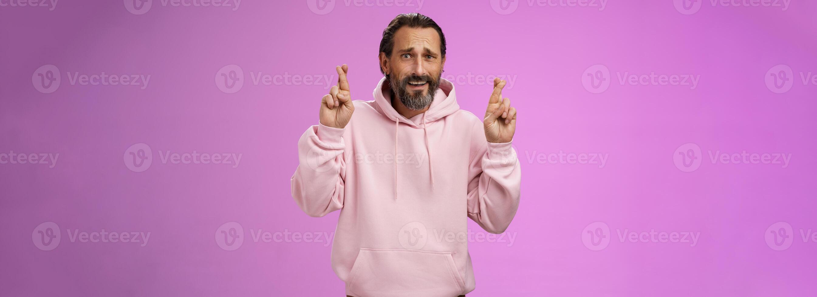 Nervous unsure hopeful handsome bearded 40s man in pink stylish hoodie cringing worried cross figers good luck make wish hope not gonna lose job supplicating praying dream come true photo