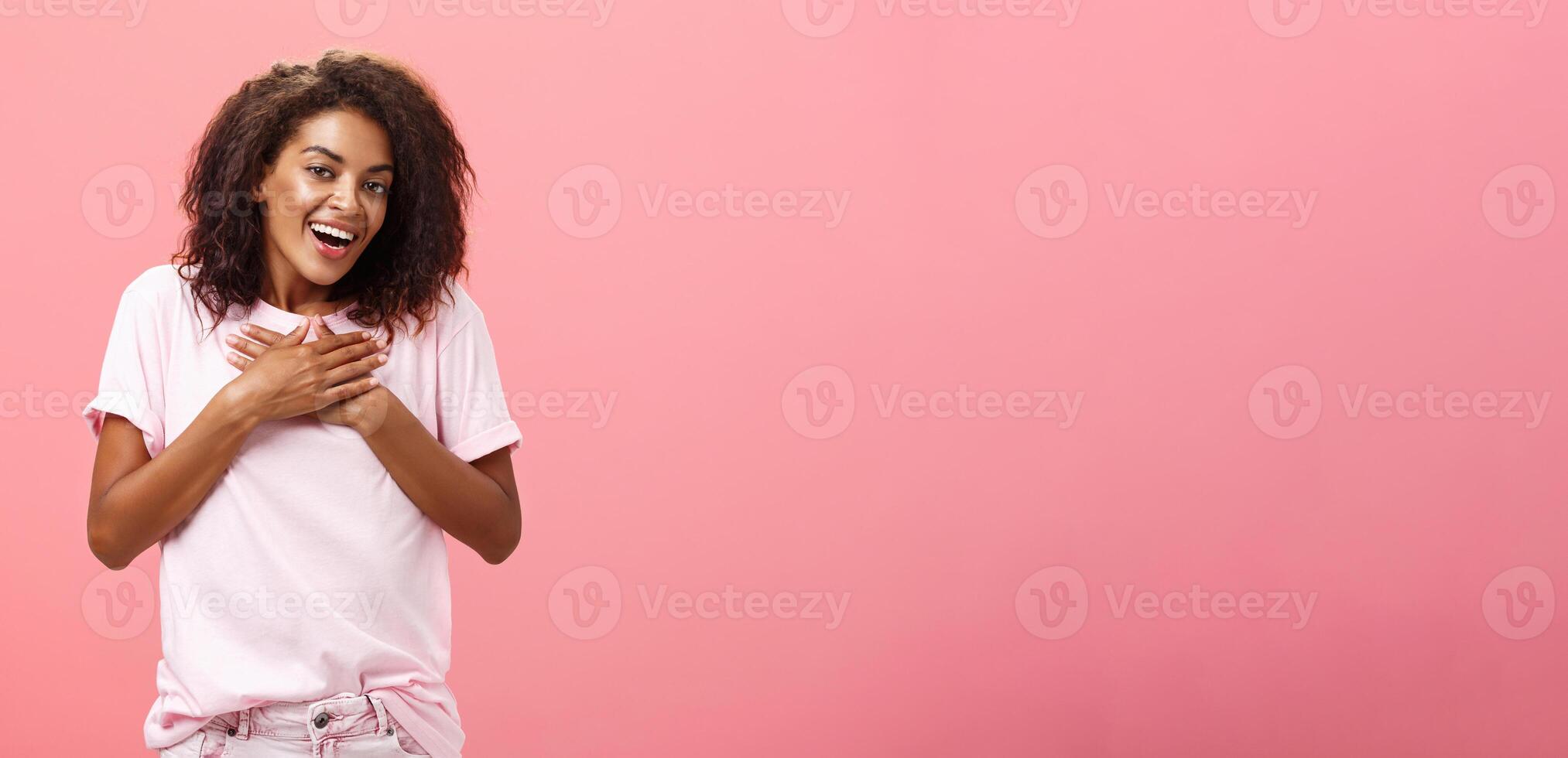 Portrait of charming delighted african american woman with curly haircut holding palms on heart pleased and grateful thanking friend for help smiling happily and thankful at camera over pink wall photo