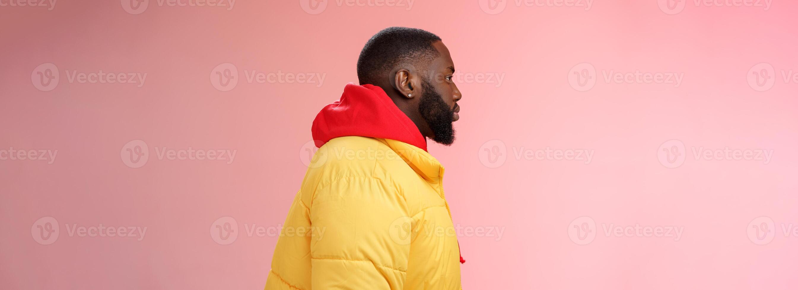 Profile studio shot bearded young 25s african guy in yellow jacket red hoodie look left normal unbothered relaxed expression standing queue order fastfood pink background, waiting take-away photo