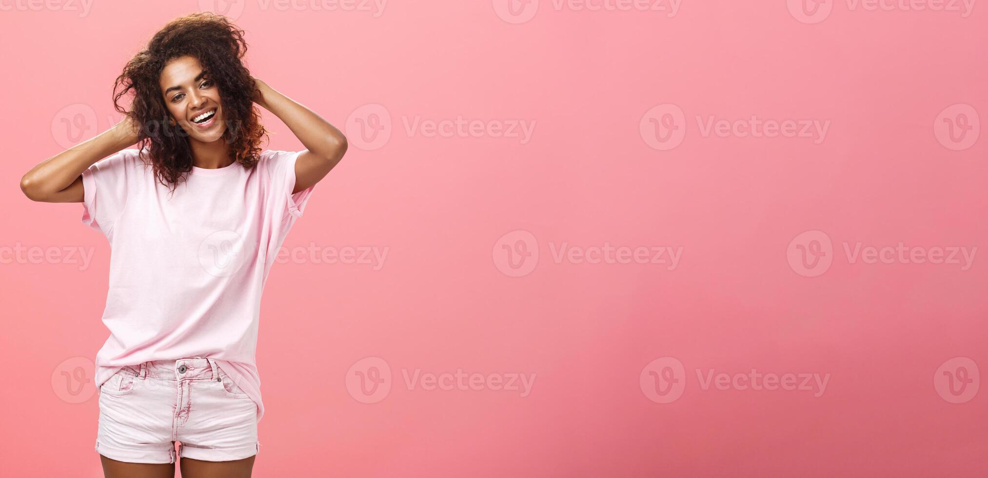 Girl enjoying nice weather chilling making photos for social network touching curly hair gently and carefree smiling broadly at camera standing over pink background happy and chill