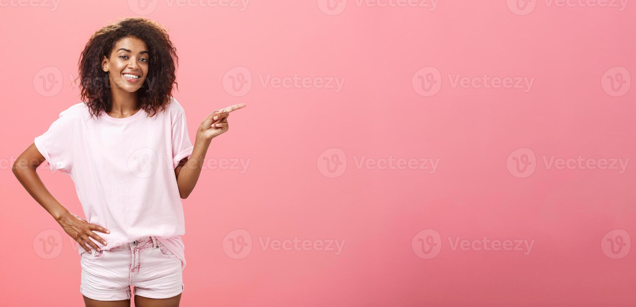 Hey look. Portrait of cute chill and friendly stylish african american woman with afro hairstyle holding hand on waist pointing right and smiling joyfully over pink background photo