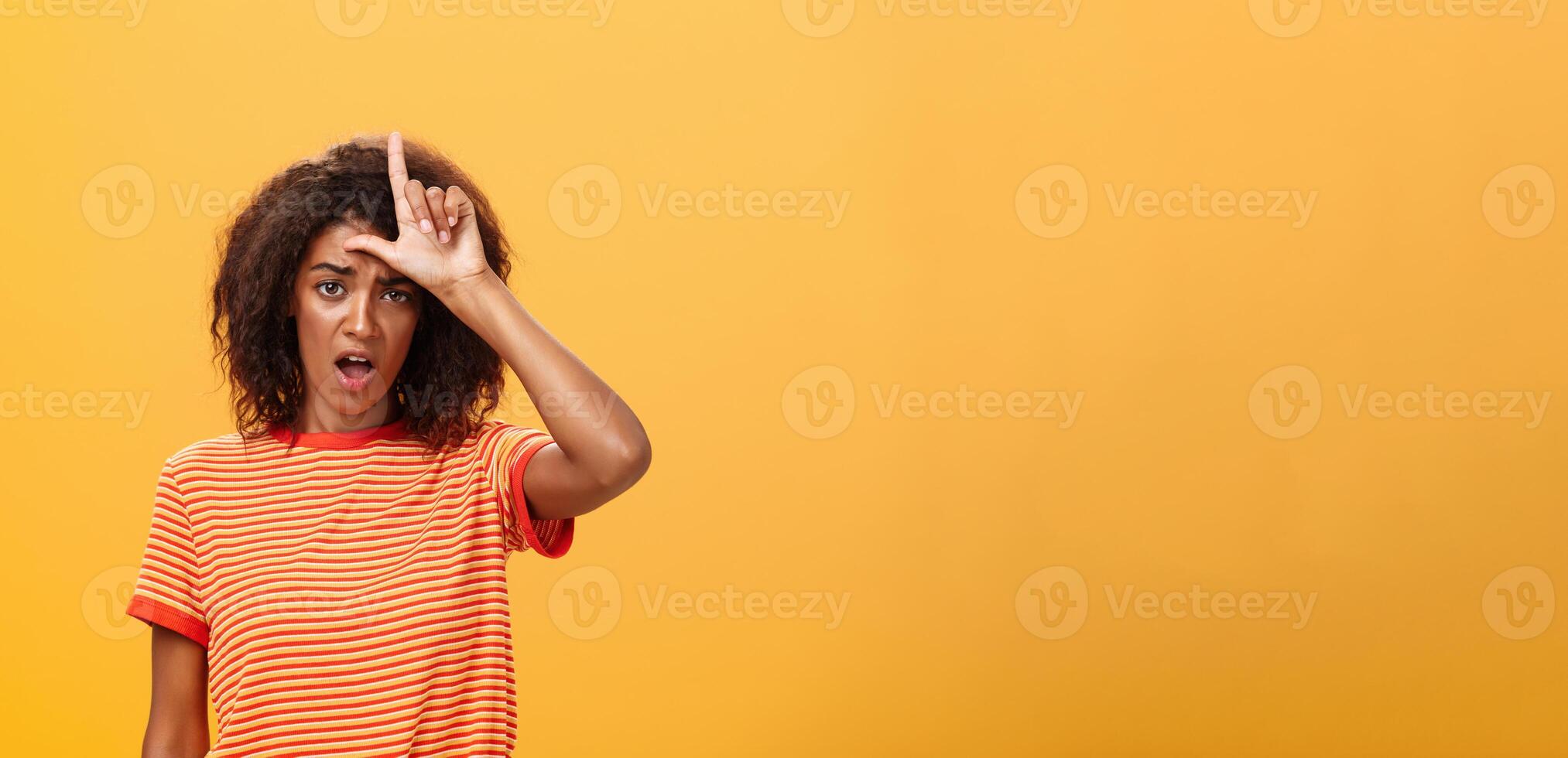 Girl thinks she loser. Portrait of gloomy bothered and displeased african american woman with afro hairstyle showing l word over forehead complaining feeling gloomy and unhappy over orange background photo