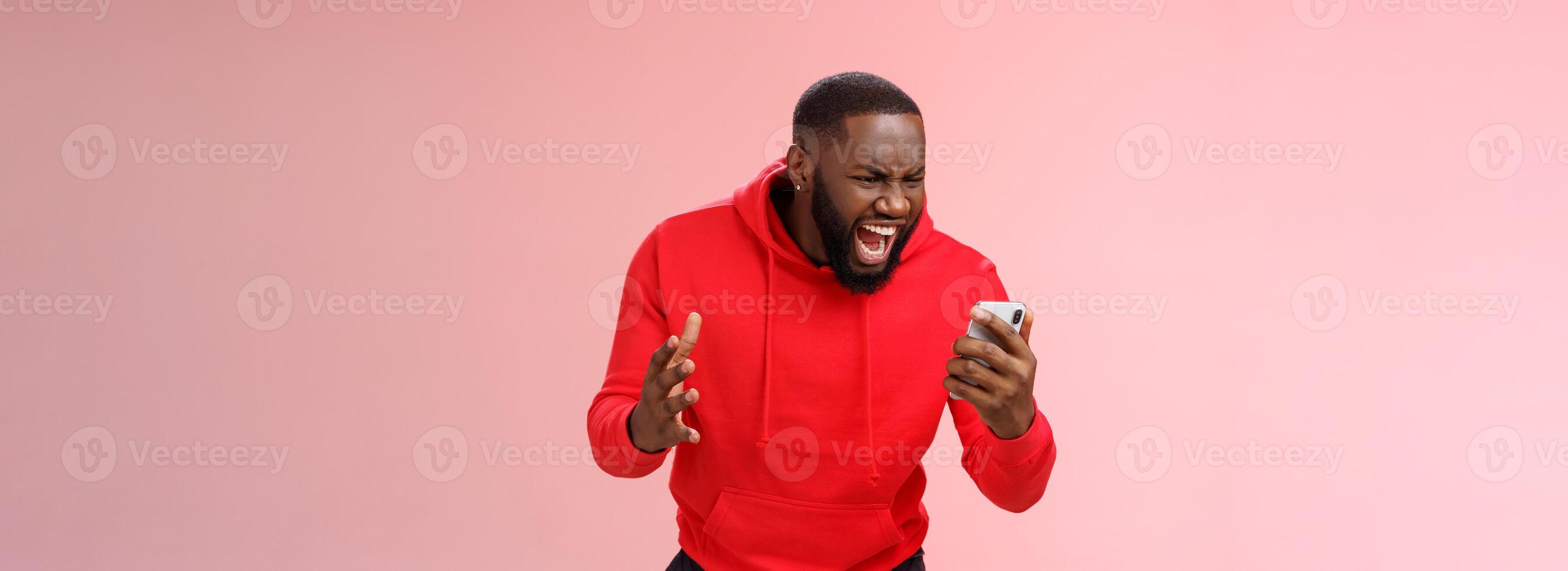 Angry furious african-american bearded guy lose control emotions shouting outraged annoyed smartphone display look phone insane stooping frowning rage, standing irritated pink background photo