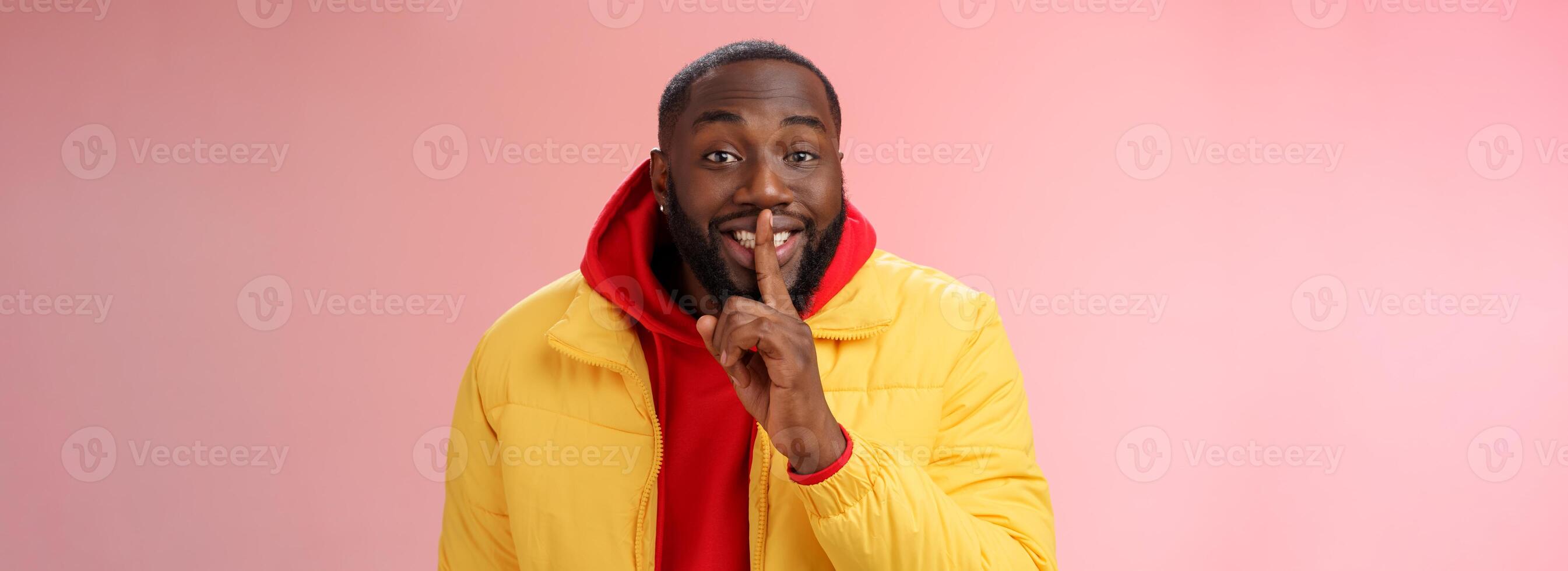 Charming happy young african-american bearded guy in yellow jacket red hoodie prepare lovely birthday surprise smiling show shush shh gesture index finger on mouth promise keep secret photo