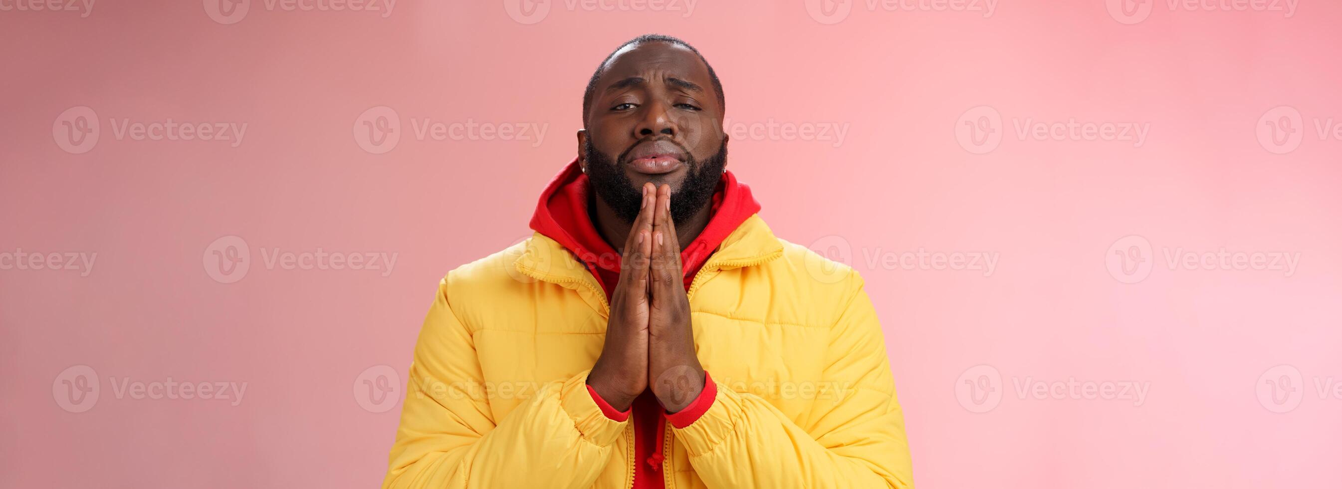 Miserable cute african-american bearded man in yellow coat asking help begging press palms supplicating apologizing please help, standing pink background sad need advice lending money photo