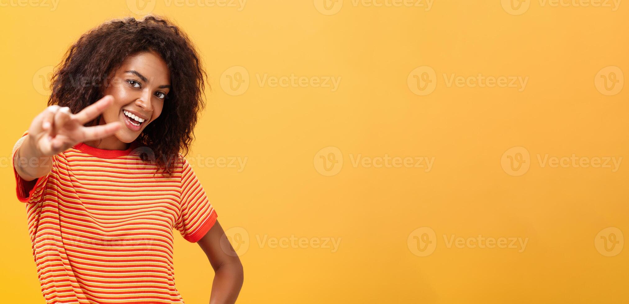 Hey peace my friend. Charming outgoing and confident carefree dark-skinned girl with afro hairstyle in trendy t-shirt pulling hand with victory gesture towards camera smiling over orange wall photo