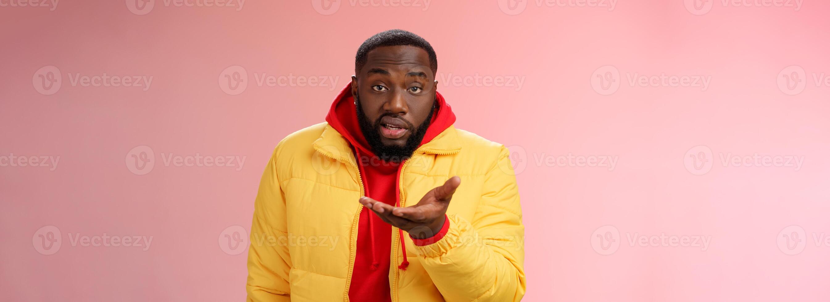 Bothered rude ignorant african-american bearded man pointing palm camera look dumb perplexed, standing confused cannot get clue what happening, wearing yellow jacket, pink background photo