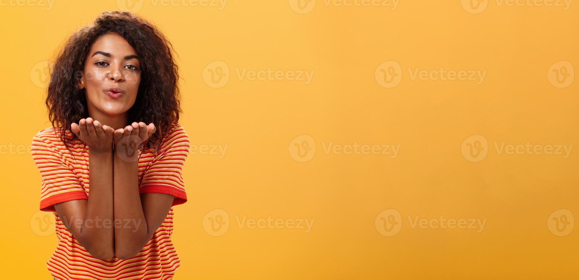 Sending passionate kiss to most loving person. Romantic attractive and stylish young african american girlfriend with curly hairstyle bending towards camera with slight smile, folded lips blowing mwah photo