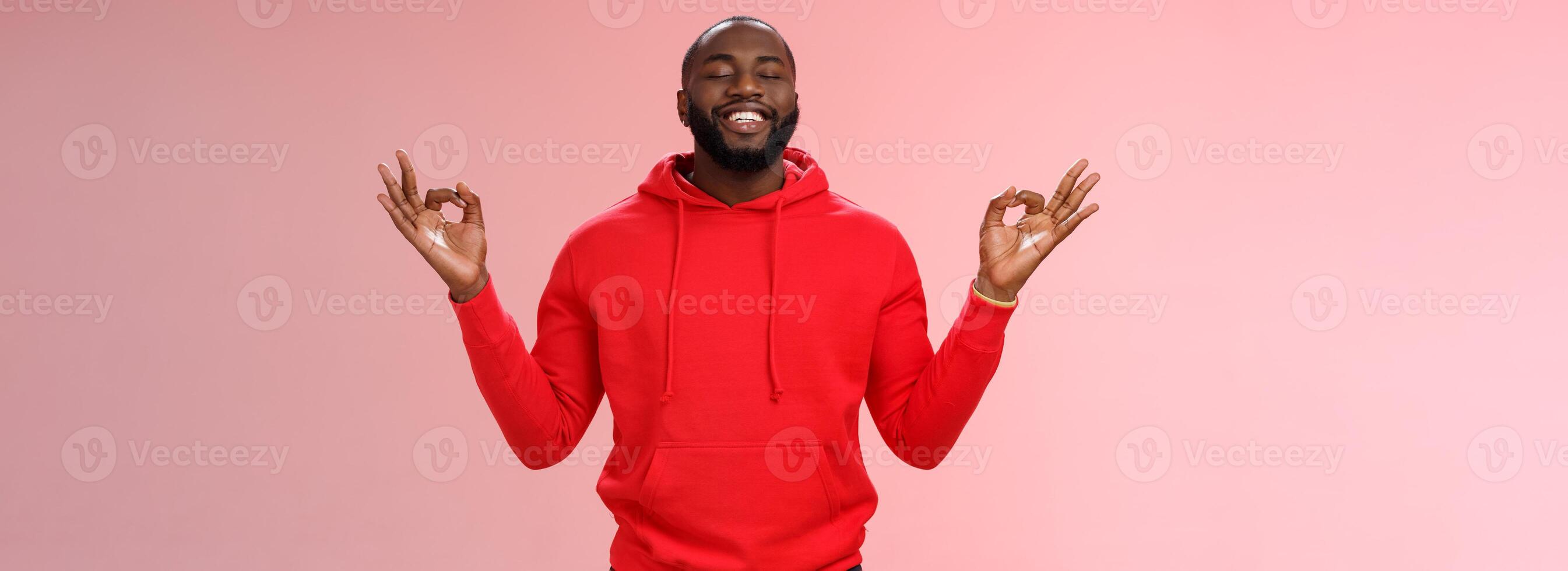 Happy african american bearded man in red hoodie meditating found peace nirvana smiling delighted close eyes relaxed relieved standing lotus mudra pose search zen, practice yoga, pink background photo