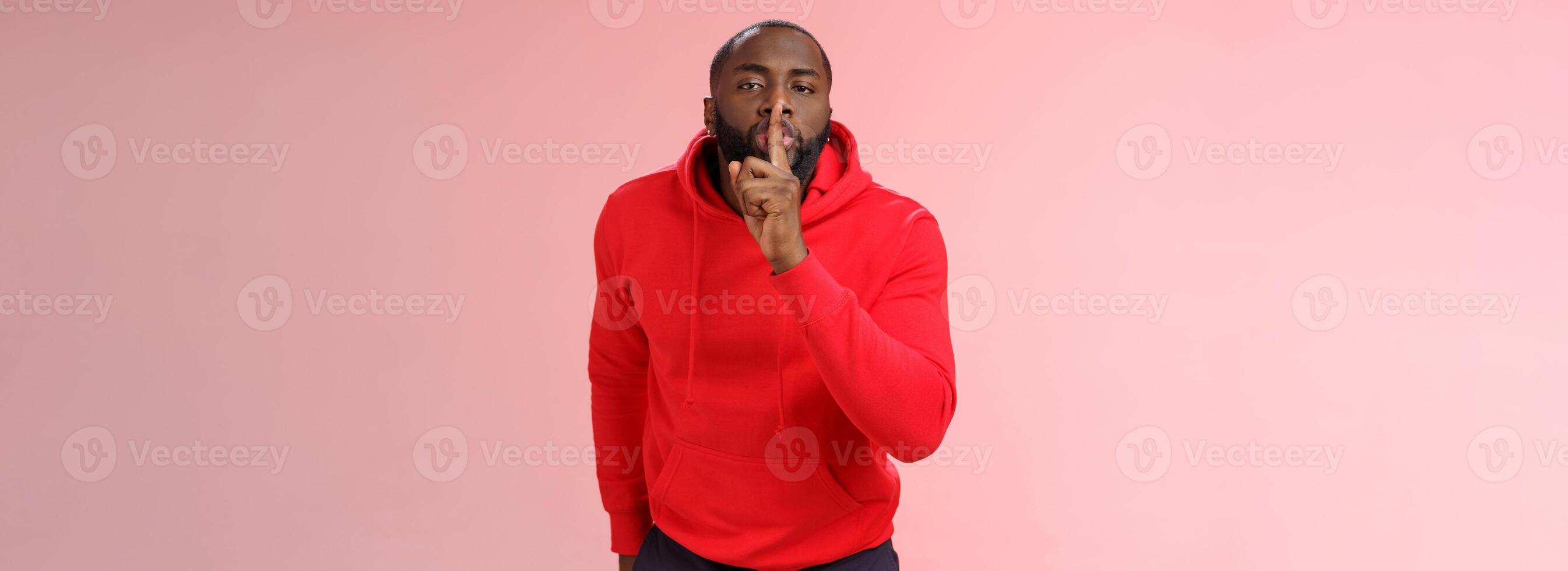 Shhh it taboo. Portrait sensual funny cute african-american bearded man in red hoodie bending camera show shush gesture index finger on lips telling secret asking keep quiet not tell anyone mystery photo