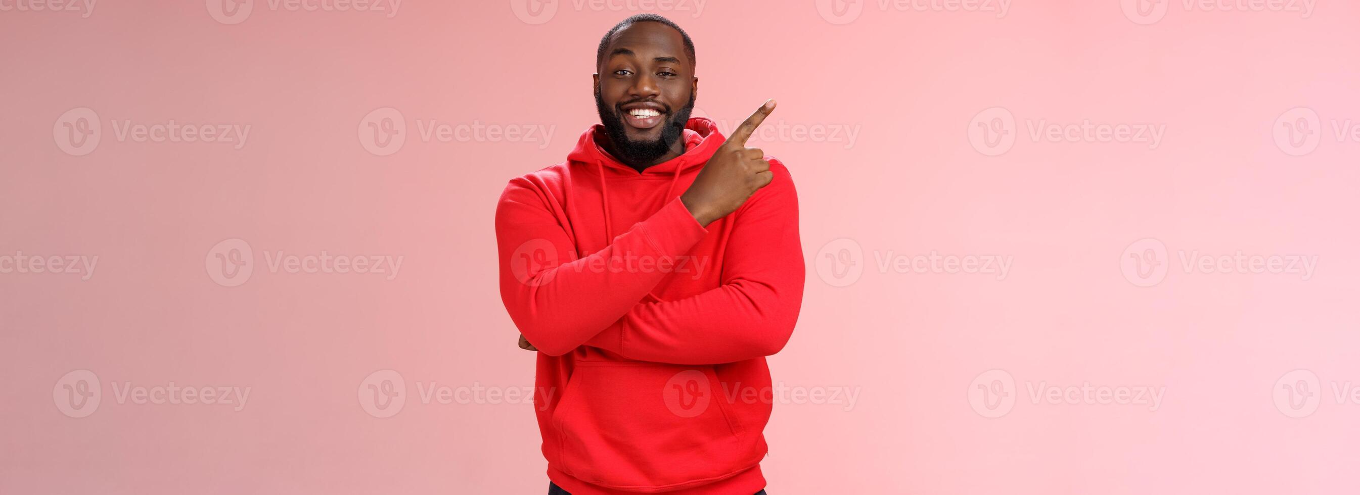 Cheeky outgoing smiling attractive african-american male model in red hoodie grinning energized pointing upper left corner indicating awesome product recommend promo, standing pink background photo